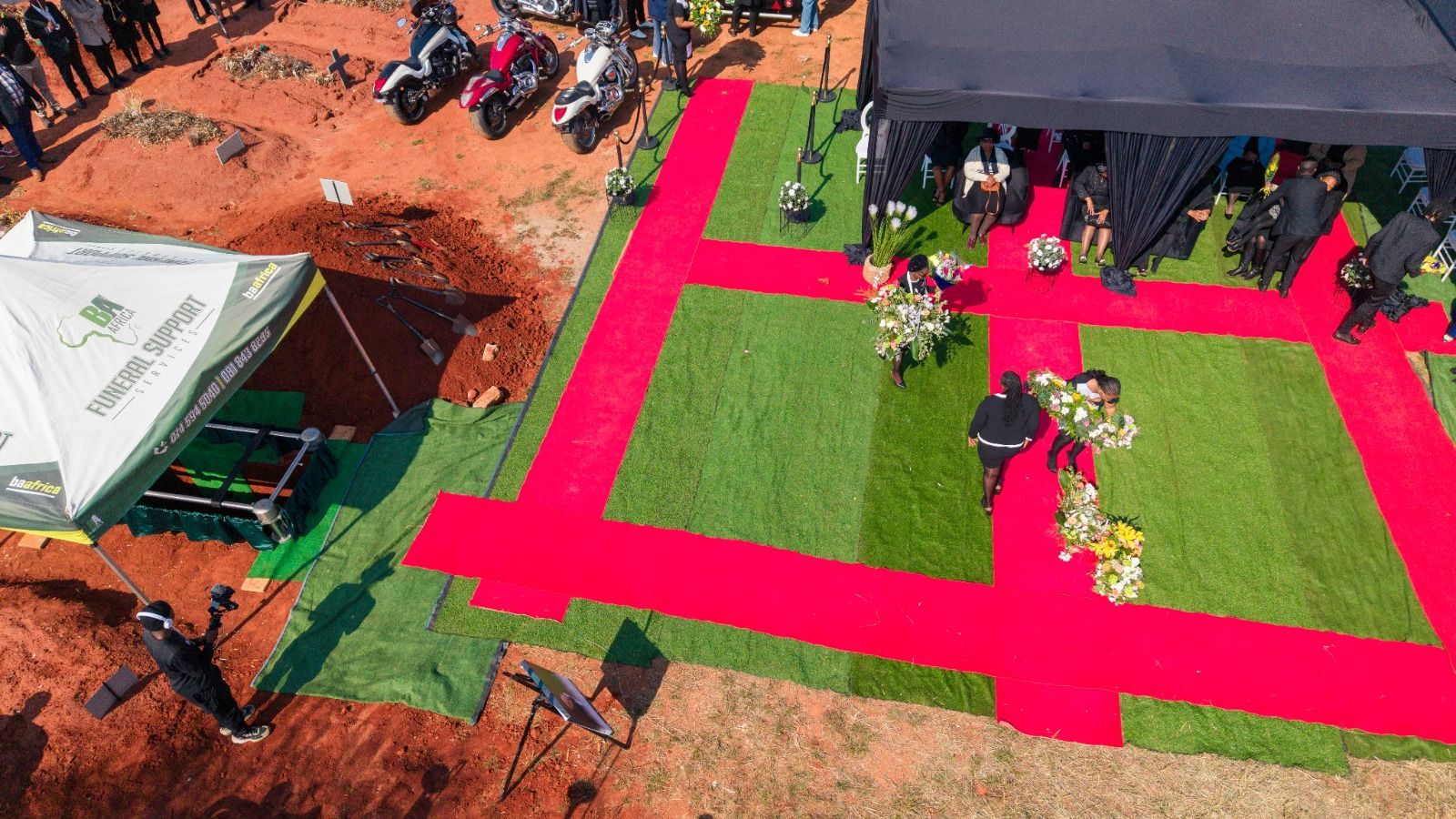 Aerial view of a BaAfrica graveside: red carpet laid in a cross over green turf, black draped gazebo and the open grave