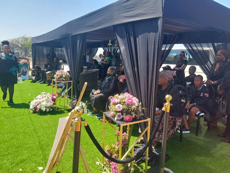 A black draped gazebo with gold stanchions and floral arrangements, family seated for the service